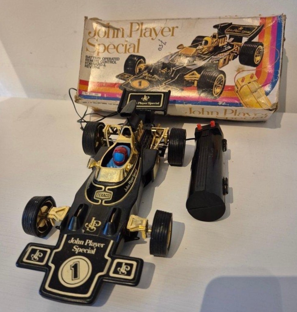 A vintage toy race car, styled as the John Player Special, with a black and gold design, featuring a driver figure and a battery pack, displayed alongside its original packaging.